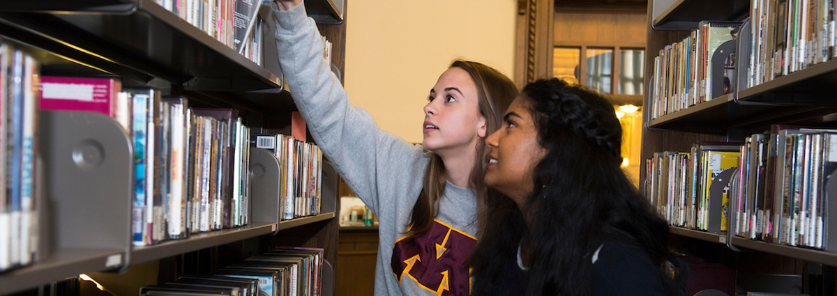 A student pulls a book off a shelf next to another student.