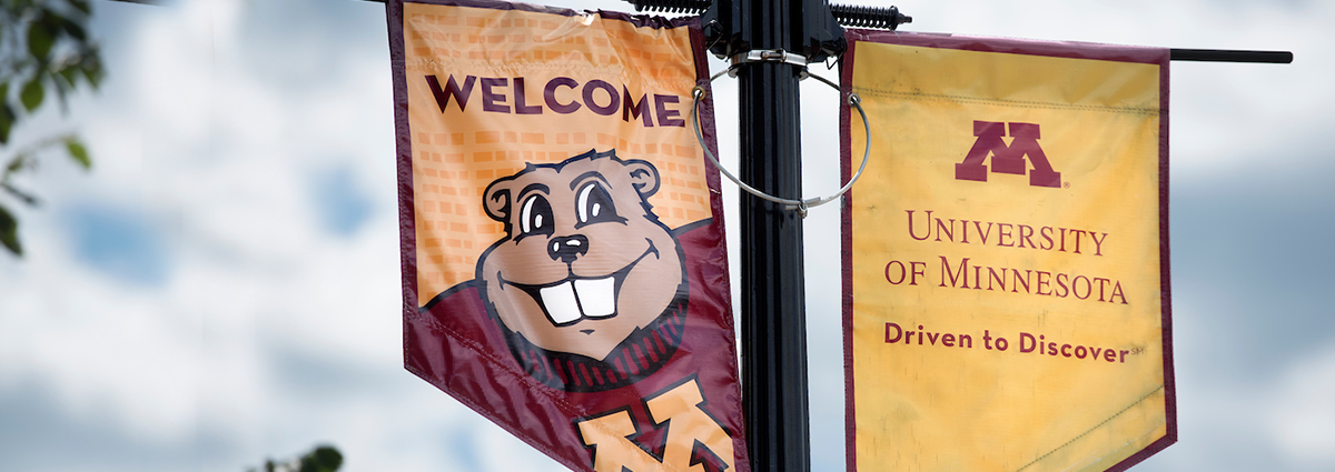 A close up of two banners hanging from a lamp post. One says "welcome" and the other features the UMN logo.