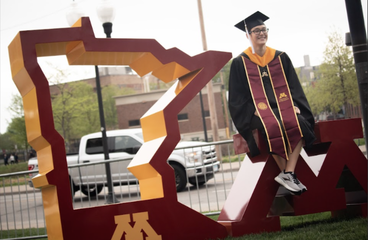 a person in a cap and gown sitting on a UMN logo sculpture